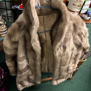 May include: A vintage faux fur coat in shades of brown and tan hangs on a blue hanger. The coat has a wide collar and a silky, light brown lining. The coat is draped over a wooden chair.