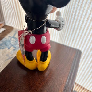 Puede incluir: Una figura de Mickey Mouse, vista desde atr&aacute;s, con orejas y cabeza negras, pantalones cortos rojos con botones blancos, zapatos amarillos y guantes blancos. Una etiqueta est&aacute; adjunta con el logotipo de Disney. La figura est&aacute; sobre una superficie marr&oacute;n oscuro.