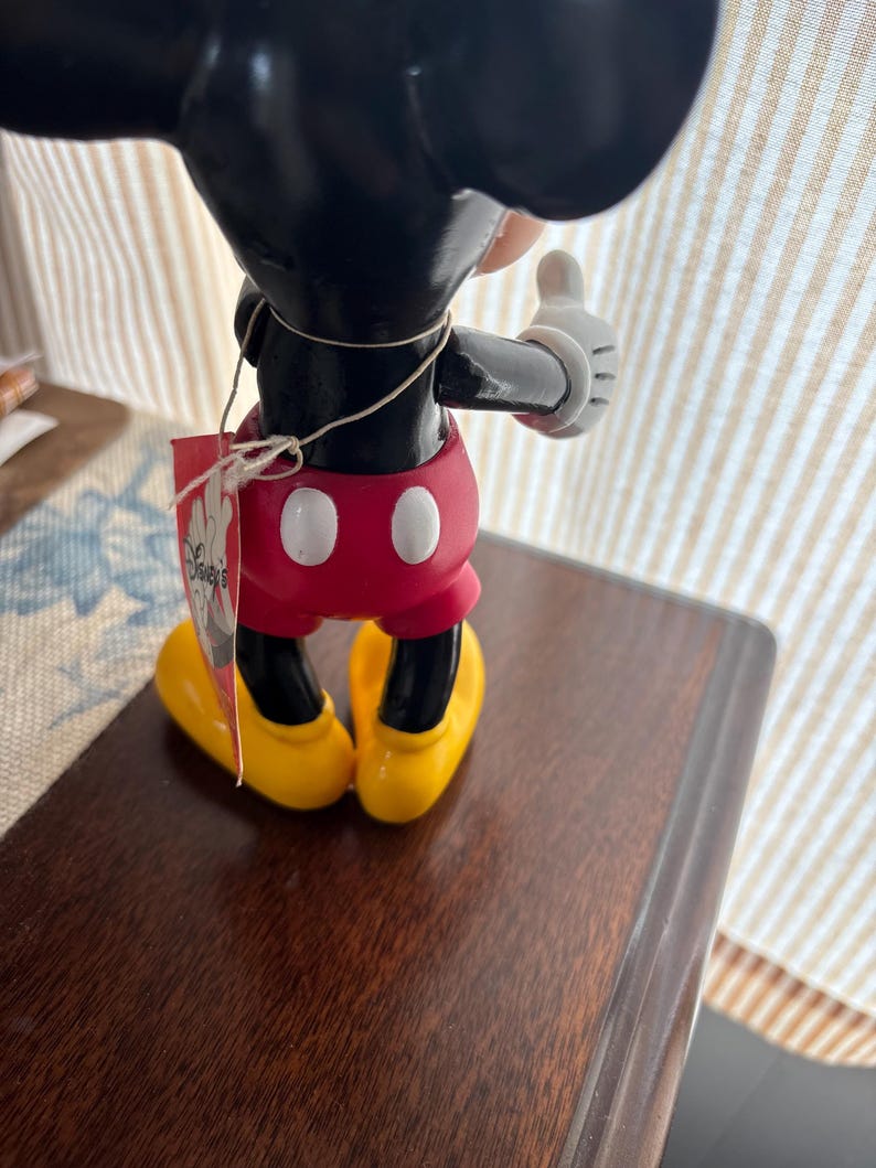 Puede incluir: Una figura de Mickey Mouse, vista desde atr&aacute;s, con orejas y cabeza negras, pantalones cortos rojos con botones blancos, zapatos amarillos y guantes blancos. Una etiqueta est&aacute; adjunta con el logotipo de Disney. La figura est&aacute; sobre una superficie marr&oacute;n oscuro.