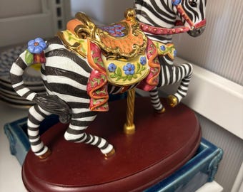 Può includere: Statuetta decorativa a forma di zebra con strisce bianche e nere, una sella colorata e una briglia rossa. La statuetta è montata su una base ovale rosso scuro e poggia su un vassoio di ceramica blu. La zebra ha un palo dorato.