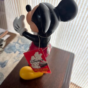 Puede incluir: Una figura de Mickey Mouse con orejas y cabeza negras, cara color melocot&oacute;n y guantes blancos. La figura lleva pantalones cortos rojos y zapatos amarillos. Una etiqueta roja con el logo de Disney est&aacute; adjunta.