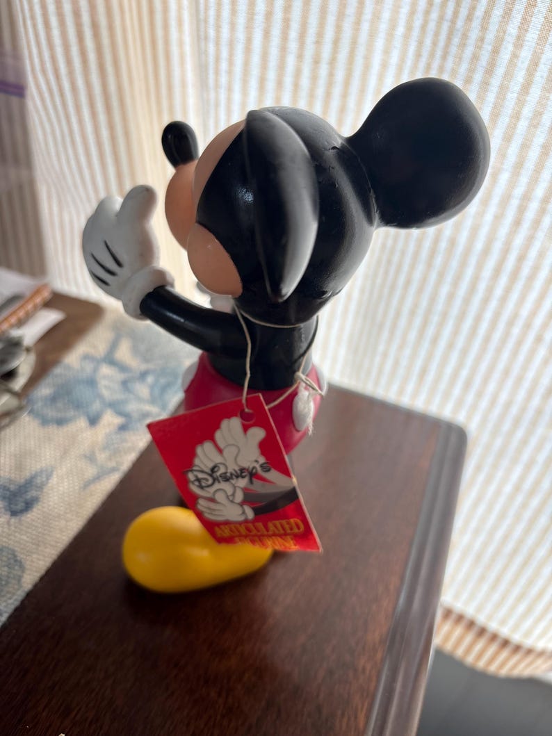Puede incluir: Una figura de Mickey Mouse con orejas y cabeza negras, cara color melocot&oacute;n y guantes blancos. La figura lleva pantalones cortos rojos y zapatos amarillos. Una etiqueta roja con el logo de Disney est&aacute; adjunta.