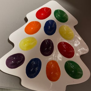 May include: A white ceramic Christmas tree-shaped paint palette with 12 individual wells filled with different colors of paint, including red, orange, yellow, green, blue, purple, and black.