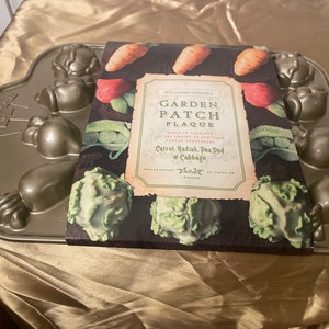 May include: A brown metal baking pan with a garden patch theme. The pan has molds for carrots, radishes, pea pods, and cabbages. The pan is next to a cookbook titled "Garden Patch Plaque" by William Sonoma.