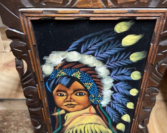 May include: A framed black velvet painting of a Native American person wearing a colorful headdress. The painting is in a dark wood frame with intricate carvings. The painting is signed "Little Big Chief".