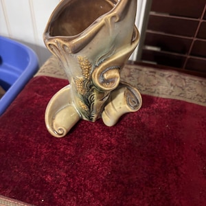 May include: A ceramic vase with a unique, asymmetrical design. The vase features a light beige base with brown and green accents, and a textured surface. Decorative floral elements are visible on the front. The vase is likely for decorative purposes.