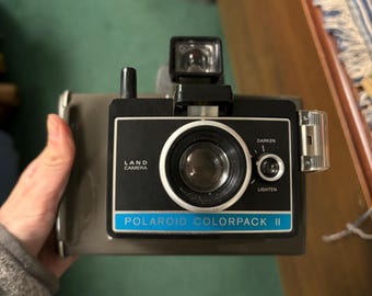 May include: A black Polaroid Colorpack II instant camera with a silver trim and a blue label. The camera has a lens, a flash, and a light/dark adjustment dial.