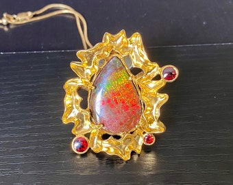 May include: A gold pendant necklace with a teardrop-shaped iridescent gemstone surrounded by a textured gold frame. Three small red gemstones are set in the frame.