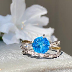 May include: A silver ring with a large blue topaz gemstone and a smaller band of diamonds. The ring has a gold accent on the band.