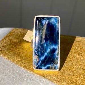 May include: A silver ring with a large, rectangular blue tiger's eye gemstone. The stone has a swirling pattern of blue and white.