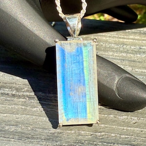 May include: A rectangular pendant with a silver chain. The pendant features a large, clear, rectangular stone with blue, yellow, and white iridescent colors. The stone is set in a silver frame.
