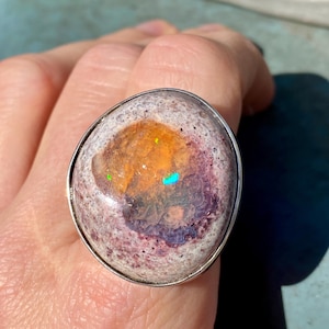 Könnte beinhalten: Ein ovaler Ring mit einem farbenfrohen Opalstein. Der Opal zeigt Farbtöne von Orange, Lila und Weiß mit schillernden grünen und blauen Blitzen. Der Stein ist in einem silberfarbenen Metallband gefasst, was ein auffälliges Schmuckstück ergibt.