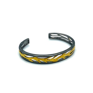 May include: A black and gold bangle bracelet with a delicate, branch-like design. The bracelet is made of metal and has a polished finish.