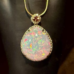 Könnte beinhalten: Eine goldene Halskette mit einem großen, birnenförmigen Opalanhänger. Der Opal zeigt schillernde Farben und ist von kleinen, klaren Steinen umgeben. Ein kleinerer, quadratischer Stein sitzt über dem Opal und ist mit einer zarten Goldkette verbunden.
