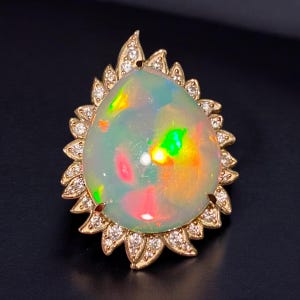 Könnte beinhalten: Ein Ring aus Perlmutt-Opal, der in Gold gefasst ist und einen Heiligenschein aus kleinen Diamanten hat. Der Opal zeigt einen Regenbogen aus Farben, darunter Grün, Rot und Gelb.