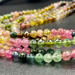 Puede incluir: Una hebra de cuentas de piedras preciosas facetadas en tonos rosa, verde y marrón. Las cuentas están dispuestas en un patrón de arcoíris, con las cuentas rosas en un extremo y las cuentas marrones en el otro.