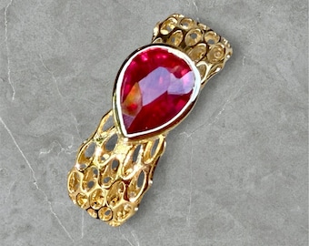 May include: A gold-toned brooch featuring a large, teardrop-shaped, red gemstone. The gemstone is set in a gold frame and is surrounded by an openwork design. The brooch has a textured, openwork design on the sides.