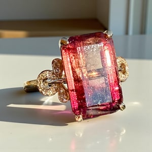 May include: A gold ring featuring a large, rectangular, pink gemstone. The gemstone has a gradient of colors, from deep red to a lighter pink. The ring has a floral design with small, clear stones on the sides.