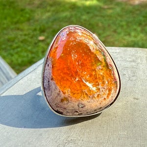 Könnte beinhalten: Ein großer, tropfenförmiger Ring mit einem leuchtenden, mehrfarbigen Stein. Der Stein weist Farbtöne von Orange, Gelb und Lila auf und ist in einem silbernen Band gefasst. Der Ring wird auf einer grauen Oberfläche mit grünem Hintergrund präsentiert.