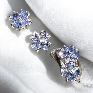 May include: A jewellery set consisting of two stud earrings and one ring, each featuring a four-petaled flower design. The pieces are crafted from silver-toned metal and set with light blue, pear-shaped gemstones arranged in a floral cluster.