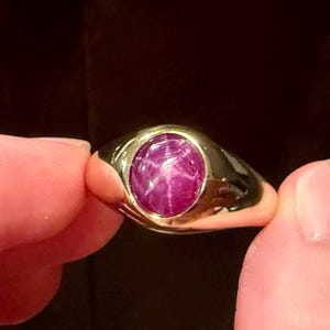 May include: A gold ring featuring a large, oval-shaped, star ruby gemstone. The ruby is a deep pinkish-red color, and the gold band has a smooth, polished finish. The ring is held between two fingers.
