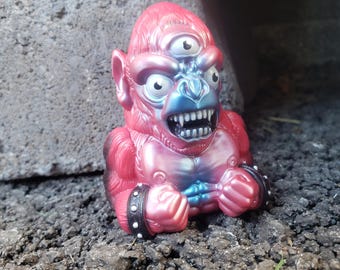 Può includere: Una statuetta in vinile stilizzata di un gorilla a tre occhi con una finitura sfumata metallizzata rosa e blu. La creatura presenta denti bianchi in vista, una fronte prominente e polsini neri borchiati sui pugni chiusi.