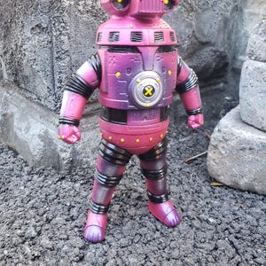 May include: A purple and black robot action figure with a rounded head, yellow eyes, and a circular chest emblem featuring a yellow X. The figure has segmented limbs, a metallic finish, and a sturdy build with visible mechanical details.