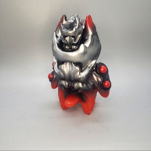 May include: A red and silver toy figure with a detailed head and multiple red accents. The figure has a menacing expression and appears to be a monster or creature.