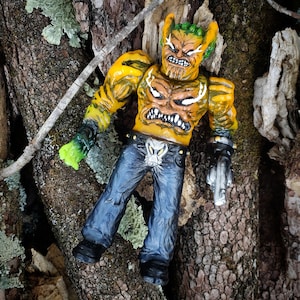 May include: A yellow and green action figure with a monster face and a gun. The figure is wearing blue jeans and a belt with a skull design.