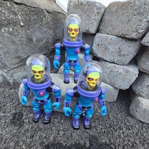 May include: Three identical action figures featuring yellow skull heads with pink eyes, encased in clear dome helmets. The figures have blue and purple suits with black shoulder accents and a small pink circular detail on the chest area.