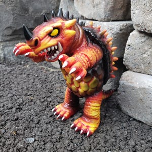 May include: A bipedal monster figurine featuring a vibrant orange and red color scheme with yellow accents. The creature has a prominent black horn on its snout, sharp white claws, yellow eyes, and a textured, scaly body with a dark shell-like back.