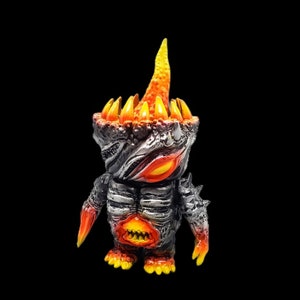 May include: A small, black and silver figurine with a fiery orange and yellow head. The figurine has a spiky crown and a large, open mouth with sharp teeth.