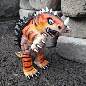May include: A stylized monster figurine featuring a vibrant orange and yellow body with black tiger-like stripes. The creature has a brown textured belly, a row of sharp black spikes along its back, white horns, and white claws on its hands and feet.
