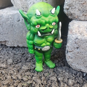 May include: A green goblin figurine with a muscular build, pointed ears, and two white horns on its head. The creature has red eyes, white fangs, and holds a small silver sword in its left hand. It wears a dark belt with a silver buckle.