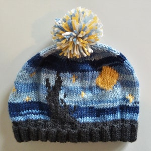 Könnte beinhalten: Eine blaue, gelbe und weiße Strickmütze mit einem Bommel oben. Die Mütze hat ein Design eines sternenklaren Nachthimmels mit einem großen gelben Mond und einer Silhouette eines Baumes.