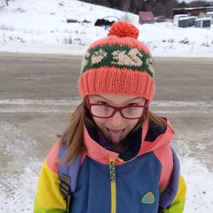May include: A young person wearing a pink knitted hat with a green and white pattern, red glasses, and a colourful jacket. The person is sticking their tongue out.