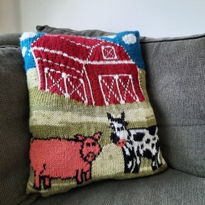 May include: A knitted pillow with a red barn, a pink pig, and a black and white cow in a green field. The pillow is on a gray couch.