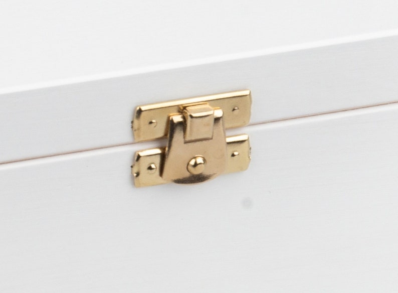 Pu&ograve; includere: Una scatola di legno bianca con una chiusura in metallo dorato. La chiusura &egrave; chiusa e ha un piccolo pomo rotondo al centro.