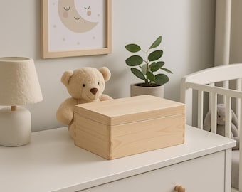 May include: A light-colored wooden box with a rectangular shape, sitting on a white dresser. A beige teddy bear is behind the box. A framed picture of a smiling moon and stars hangs on the wall. A small lamp and a potted plant are also visible.