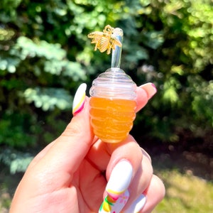 May include: A clear, honey-shaped lip gloss container filled with a golden liquid. The container has a gold-colored bee charm on top. The background is a blurred view of green foliage.