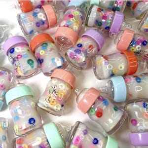 Puede incluir: Una colección de biberones en miniatura con detalles coloridos. Cada biberón tiene una tetina y tapa de color pastel, llenos de líquido transparente y pequeñas decoraciones circulares de colores. Algunos biberones tienen el texto "Gua Taylor Cosmetics".