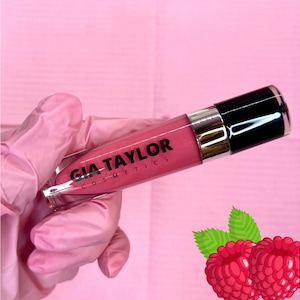 May include: A clear tube of pink lip gloss with a black cap. The tube has the text "GIA TAYLOR COSMETICS" printed on it. The lip gloss is next to two red raspberries.