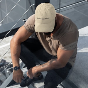 May include: A beige distressed baseball cap with the text "SATOSHI NAKAMOTO" embroidered in black.