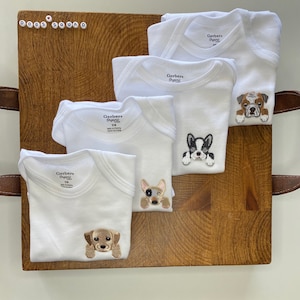Puede incluir: Cuatro bodies blancos Gerber Organic para beb&eacute; con diferentes razas de perros bordadas. Los bodies est&aacute;n dispuestos sobre una superficie de madera. Los perros son un bulldog, un bulldog franc&eacute;s, un chihuahua y un labrador.