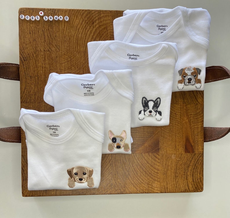 Puede incluir: Cuatro bodies blancos Gerber Organic para beb&eacute; con diferentes razas de perros bordadas. Los bodies est&aacute;n dispuestos sobre una superficie de madera. Los perros son un bulldog, un bulldog franc&eacute;s, un chihuahua y un labrador.