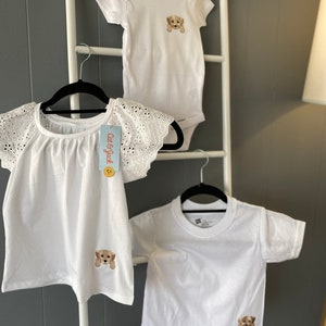 Puede incluir: Tres camisas blancas con una linda cara de cachorro bordada en cada una. Las camisas est&aacute;n colgadas en una escalera blanca con una pared gris en el fondo. Las camisas son de diferentes tama&ntilde;os y estilos, incluyendo un body de manga corta, una camiseta de manga corta y una blusa de manga corta con encaje.