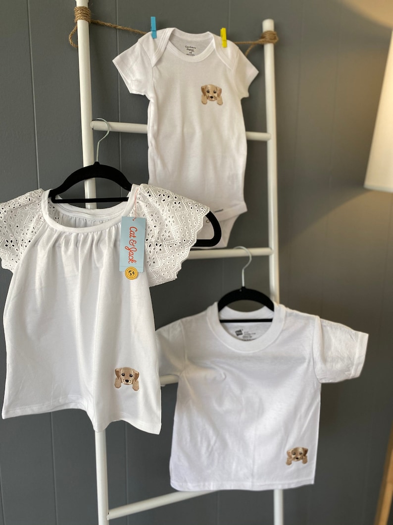 Puede incluir: Tres camisas blancas con una linda cara de cachorro bordada en cada una. Las camisas est&aacute;n colgadas en una escalera blanca con una pared gris en el fondo. Las camisas son de diferentes tama&ntilde;os y estilos, incluyendo un body de manga corta, una camiseta de manga corta y una blusa de manga corta con encaje.