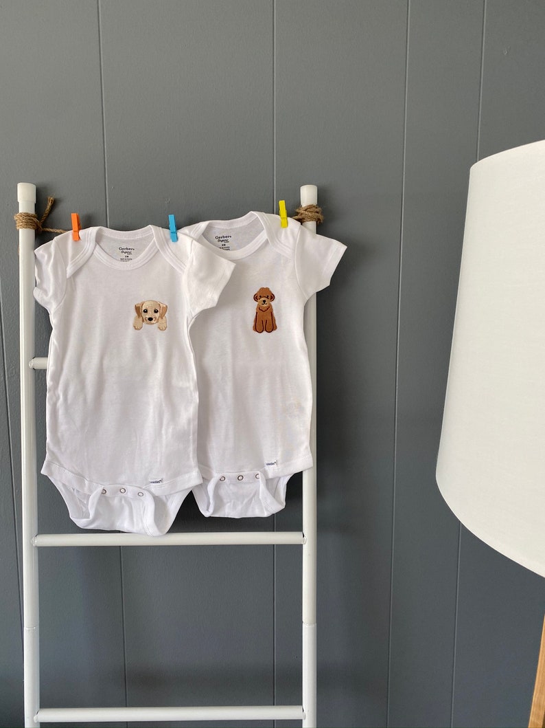 Puede incluir: Dos bodies blancos para beb&eacute; con broches. Los bodies tienen un gr&aacute;fico de un perro en la parte delantera. Un body tiene un perro marr&oacute;n y el otro tiene un perro blanco.