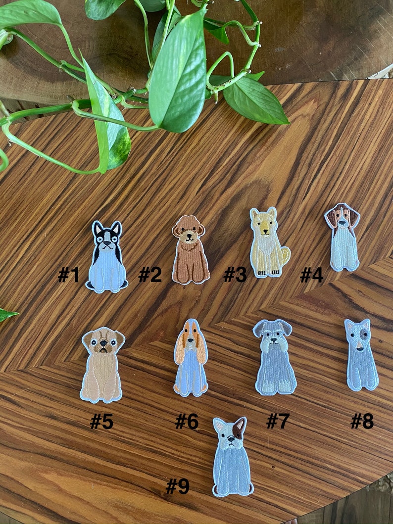 Puede incluir: Nueve parches bordados para planchar con diferentes razas de perros. Los parches est&aacute;n sobre una superficie de madera con una planta verde en el fondo.