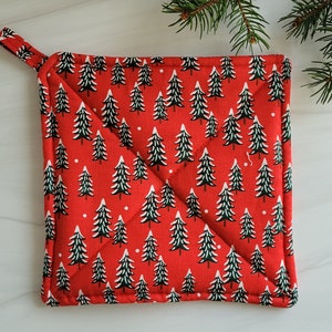 May include: A square, red Christmas potholder with a repeating pattern of green and white Christmas trees. The potholder has a small loop for hanging and is quilted with white thread. A sprig of evergreen is in the background.
